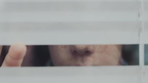 Karantän. En tonåring med fräknar kikar ut genom stängda persienner. — Stockvideo