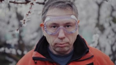 Tıbbi maskeli bir adam, gözlüklü ve çiçek açan bir ağacın arka planında özel bir takım elbiseli.