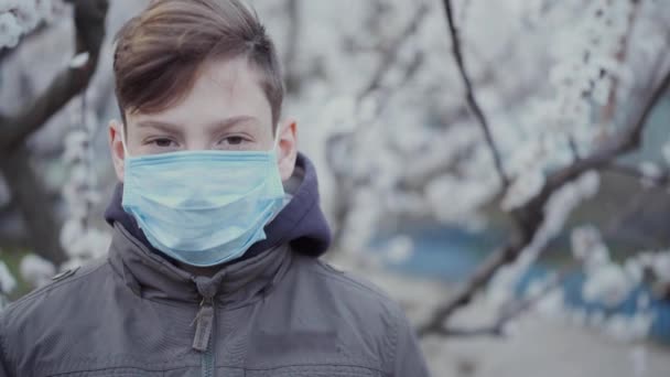 Un adolescent dans un masque médical sur un fond d'arbre en fleurs . — Video