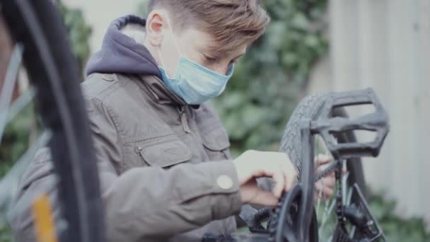 En tonåring reparerar sin cykel i en medicinsk mask. — Stockvideo
