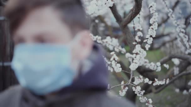 La cara de un adolescente con una máscara médica en el fondo de un árbol de primavera floreciente . — Vídeos de Stock