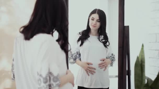 Έγκυος κορίτσι θαυμάζει τον εαυτό της και την κοιλιά της κοιτάζοντας στον καθρέφτη — Αρχείο Βίντεο