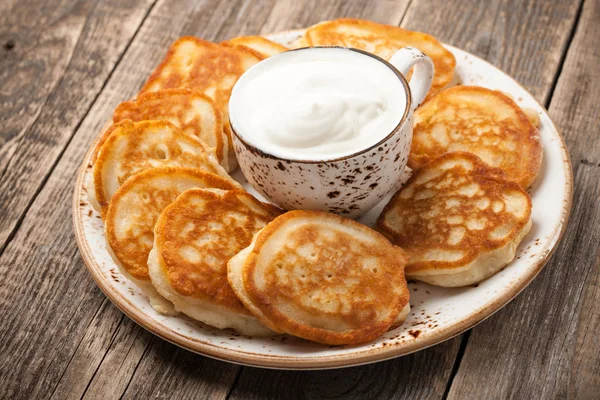 Frittelle fatte in casa e tazza di panna acida in un piatto sul tavolo di legno — Foto Stock