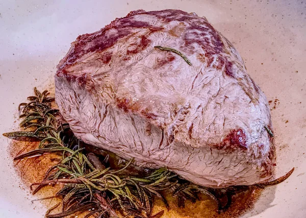 A big piece of beef meat with rosemary and olive oil cooked in a white ceramic pot. Hdr effect.