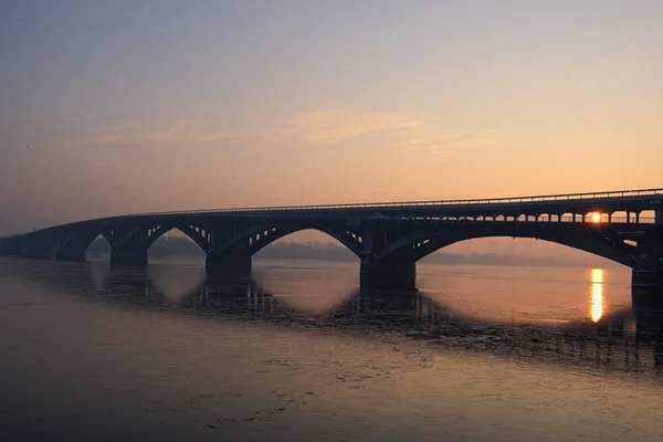 Pittoresca vista sul ponte della metropolitana (metropolitana) sul fiume Dnipro a Kiev, Ucraina. Alba al mattino d'inverno. Il disco rosso del sole fa capolino da dietro il ponte — Foto Stock