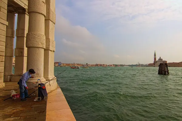 Venise, Italie-28 septembre 2019 : Un vieil homme pêche sur le remblai pavé près de Punta della Dogana. C'est un musée d'art. Paysage urbain pittoresque en arrière-plan. Venise, Italie — Photo