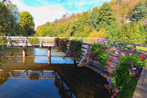 Живописный вид на старый деревянный мост, украшенный цветами на горшках. Мост отражается на поверхности воды. Солнечный осенний день. Холмы с лесом на заднем плане. Романтическая и мирная сцена. Словения — стоковое фото