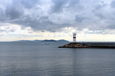 İskelenin sonundaki beyaz deniz fenerinde özel seyrüsefer ekipmanları liman girişini gösteriyor. Denizde yalnız bir balıkçı teknesi. Dramatik bulutlu gökyüzü. İstanbul, Türkiye