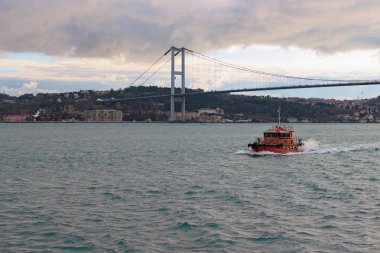 Bulutlu bir günde Boğaz 'ın (İstanbul Boğazı) manzarası. Boğaz 'da dolaşan kırmızı motorlu tekne. Arka planda Boğaz Köprüsü (15 Temmuz Şehitler Köprüsü, Birinci Köprü) var.