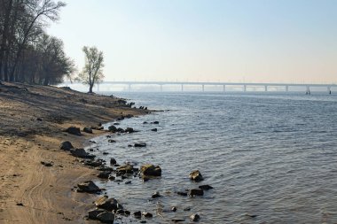Hidropark adasındaki boş plajlı Dnipro Nehri manzarası. Kıyıda bir sürü taş var. Sabah dumanında Dnipro 'nun karşısındaki Paton Köprüsü. Güneşli bir bahar sabahı. Kyiv, Ukrayna. 