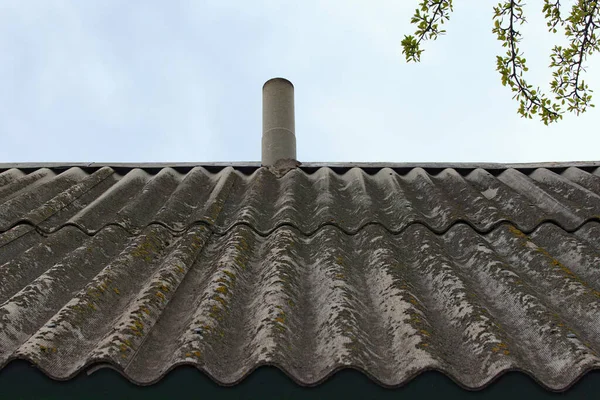 Old wavy slate roof with chimney against blue sky. Texture of old slate with moss. Shed roof covered with old asbestos sheets. Outdoor interior. Texture of old roof, slate background.