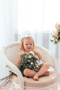 Küçük bir kız bir buket çiçekle bir sandalyede oturuyor, güneşli bir ışık..