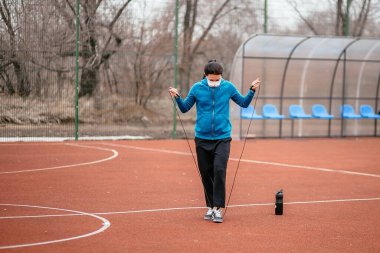 Genç bir kadın tıbbi bir maskeyle ip atlamakla meşgul. Tıbbi bir maskeyle koronavirüs enfeksiyonuna karşı koruma. Rusya 'da ilkbahar başlarında.
