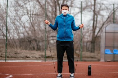 Genç bir kadın tıbbi bir maskeyle ip atlamakla meşgul. Tıbbi bir maskeyle koronavirüs enfeksiyonuna karşı koruma. Rusya 'da ilkbahar başlarında.