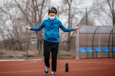 Genç bir kadın tıbbi bir maskeyle ip atlamakla meşgul. Tıbbi bir maskeyle koronavirüs enfeksiyonuna karşı koruma. Rusya 'da ilkbahar başlarında.