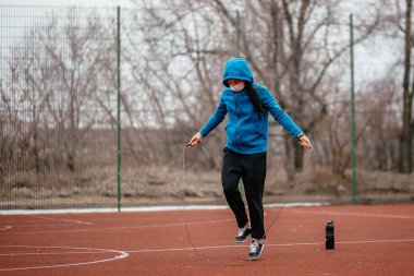 Genç bir kadın tıbbi bir maskeyle ip atlamakla meşgul. Tıbbi bir maskeyle koronavirüs enfeksiyonuna karşı koruma. Rusya 'da ilkbahar başlarında.