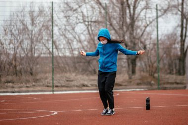 Genç bir kadın tıbbi bir maskeyle ip atlamakla meşgul. Tıbbi bir maskeyle koronavirüs enfeksiyonuna karşı koruma. Rusya 'da ilkbahar başlarında.