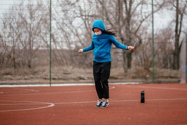 Genç bir kadın tıbbi bir maskeyle ip atlamakla meşgul. Tıbbi bir maskeyle koronavirüs enfeksiyonuna karşı koruma. Rusya 'da ilkbahar başlarında.