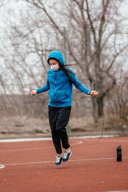 Genç bir kadın tıbbi bir maskeyle ip atlamakla meşgul. Tıbbi bir maskeyle koronavirüs enfeksiyonuna karşı koruma. Rusya 'da ilkbahar başlarında.