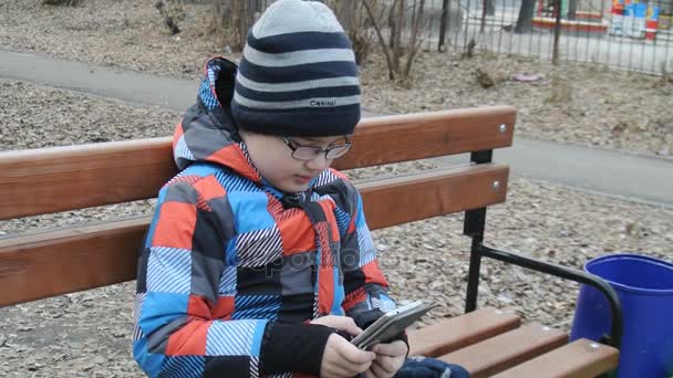 Молодой подросток играет в игру на смартфоне на скамейке снаружи . — стоковое видео