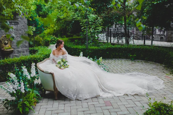 Jonge mooie bruid in luxe jurk met een lange trein zittend op een bank — Stockfoto