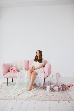 Beyaz gecelikli genç gelin stüdyodaki gül kanepesinde poz veriyor.