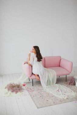 Beyaz gecelikli genç gelin stüdyodaki gül kanepesinde poz veriyor.