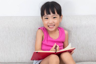 Mutlu Asyalı Çinli küçük kız kanepe yazma kitap üzerinde oturuyor