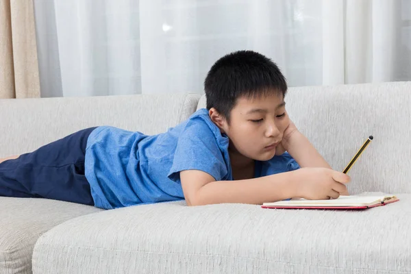 Aborrecido asiático chinês menino escrevendo livro no sofá — Fotografia de Stock