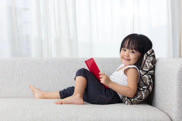 Sorridente asiatica cinese bambina lettura libro sul divano — Foto Stock