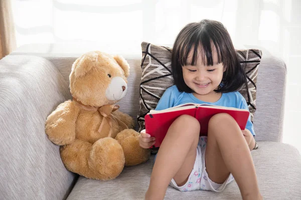 Sonriente asiático chino niña leyendo libro con osito de peluche — Foto de Stock