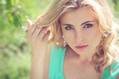 Sarışın, turkuaz elbiseli genç bir kız bahar bahçesinde yürüyor.