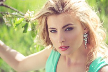 Sarışın, turkuaz elbiseli genç bir kız bahar bahçesinde yürüyor.