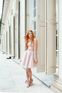 Kızıl saçlı genç kız ağaçlar ve mimari objeler arasında bir parkta yürüyor.