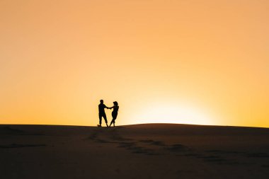 Mutlu genç bir çiftin siluetleri. Kum çölünde, turuncu günbatımının arka planında.