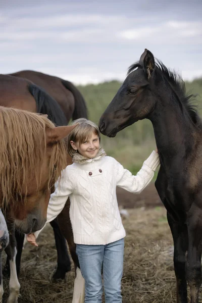 Смешной счастливый ребенок в белом свитере и джинсах стоит среди лошадей жеребцов на ферме улыбаясь. Портрет в стиле жизни — стоковое фото