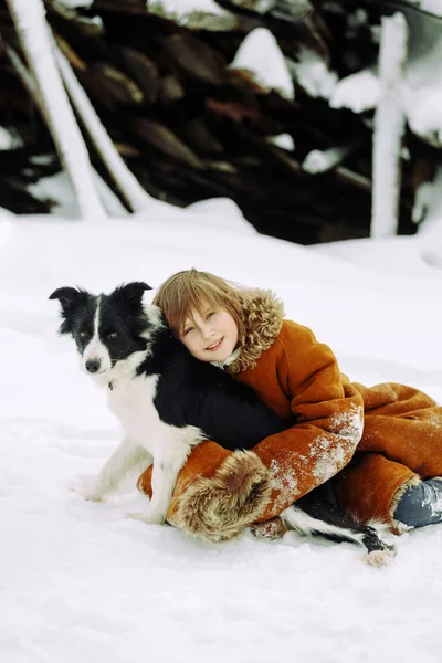 Niña sentada en la nieve en invierno con un abrigo de piel de oveja y un perro abrazador — Foto de Stock