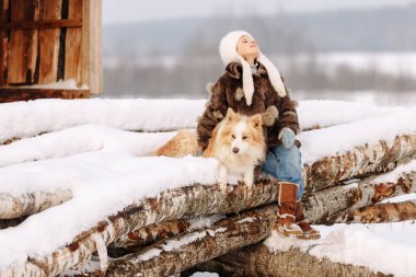 Gülümseyen küçük kız etek ve beyaz şapkası ile kışın günlükleri üzerinde oturan bir köpek