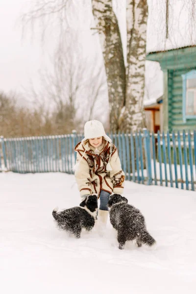 Sonriente niña linda jugando frente a una casa en invierno nieve con dos cachorros blancos y negros — Foto de Stock