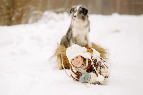 Linda niña en un abrigo de piel y botas sonriendo y tirando de un trineo en la nieve con heno en el que el perro está sentado al aire libre — Foto de Stock