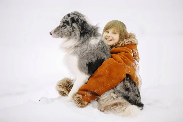 Niña sonriente en el abrigo de piel de la madre abrazando a su amigo el perro sentado en la nieve en invierno y sonriendo — Foto de Stock