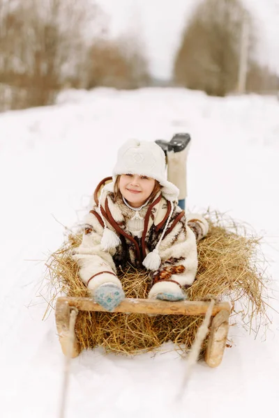 Cheerful happy smiling girl child in a fur coat and hat in the winter the snow lies on a sled with hay and looking at the camera Stock Picture