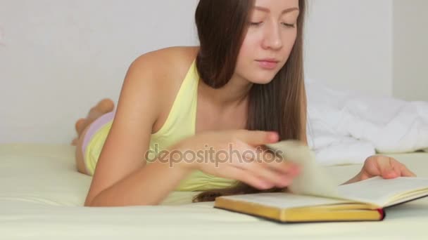 Красивая молодая девушка читает книгу лежа на кровати — стоковое видео