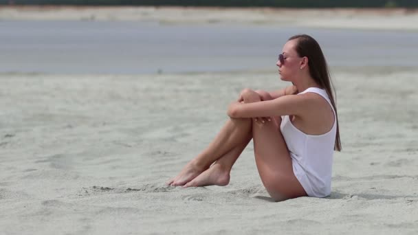 Mujer lánguida sentada con las piernas cruzadas sobre la arena tocando la piel — Vídeos de Stock