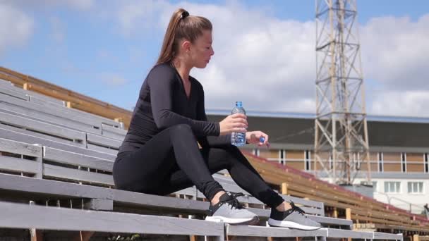 Радостная уставшая женщина в спортивной одежде отдыхает и пьет воду после тренировки на стадионе — стоковое видео