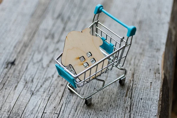 Toy wooden house with a shopping trolley on a natural wooden background. Concept photo about buying real estate in nature or outside the city.