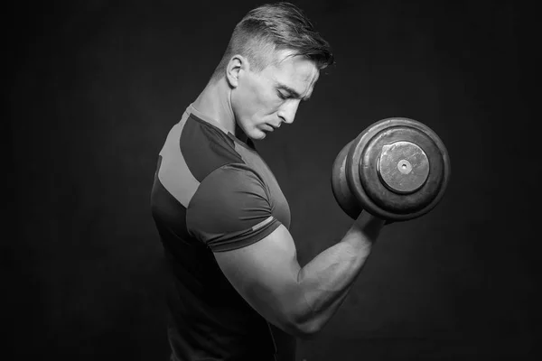 Athletic young male fitness model holds the dumbbell with light isolated on dark background.
