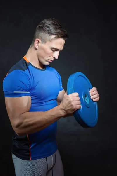 Athletic  young male fitness model holds the dumbbell with light isolated on dark background.