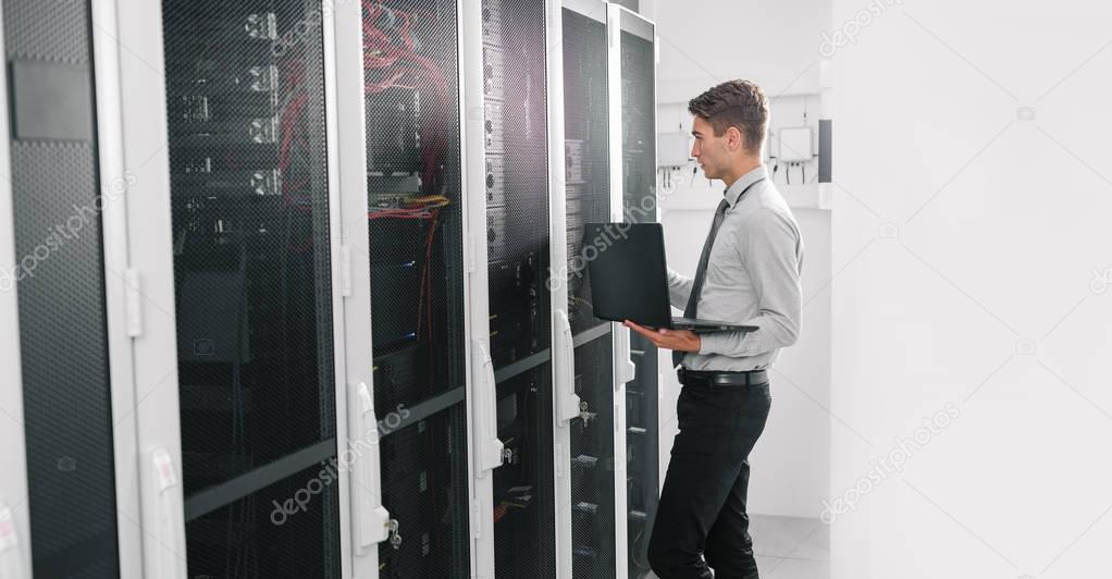 Portrait of modern young man holding laptop standing in server room working with supercomputer