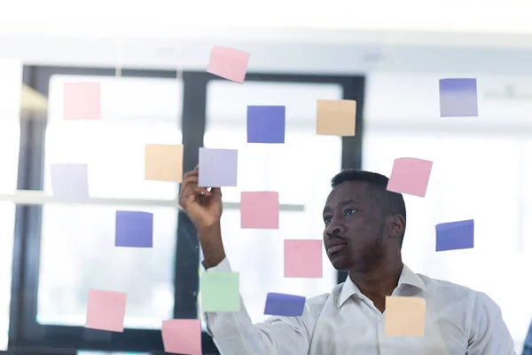 Businessman reading adhesive notes on glass wall in office and analyzing them